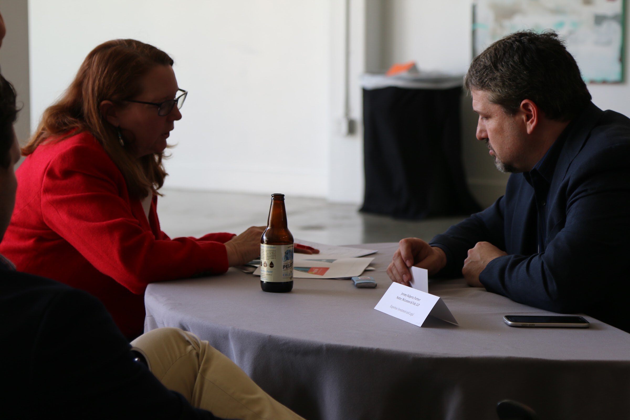 Woman in red blazer and man in navy jacket in a meeting, beer bottle and name placard on table.