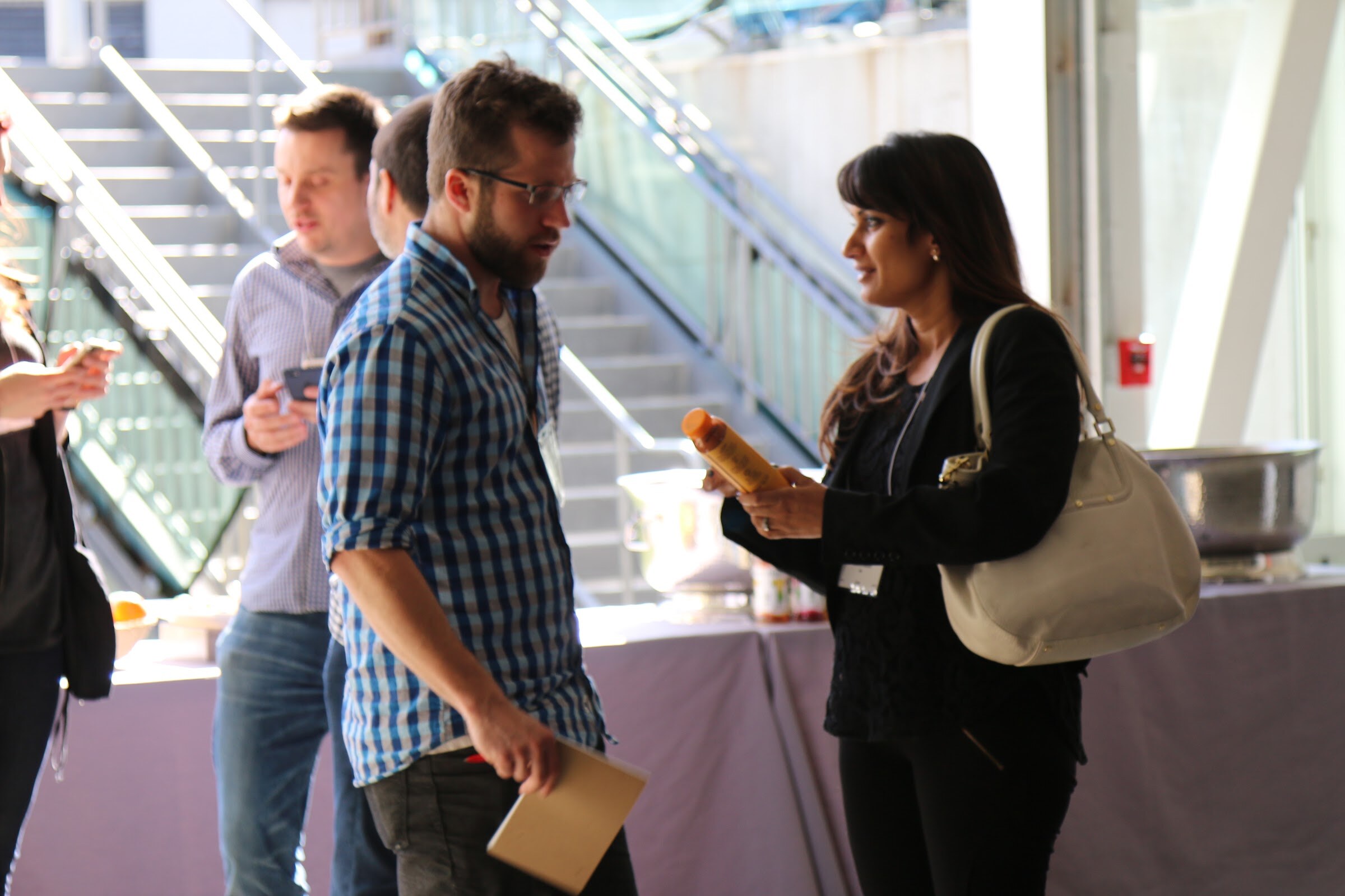 Man in plaid shirt and woman with a bag talking by buffet table before stairs.