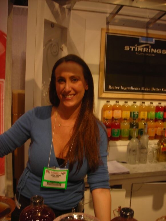 A smiling woman with long brown hair and a badge stands at a counter in front of a Stirrings sign and shelves of colorful bottled mixers.