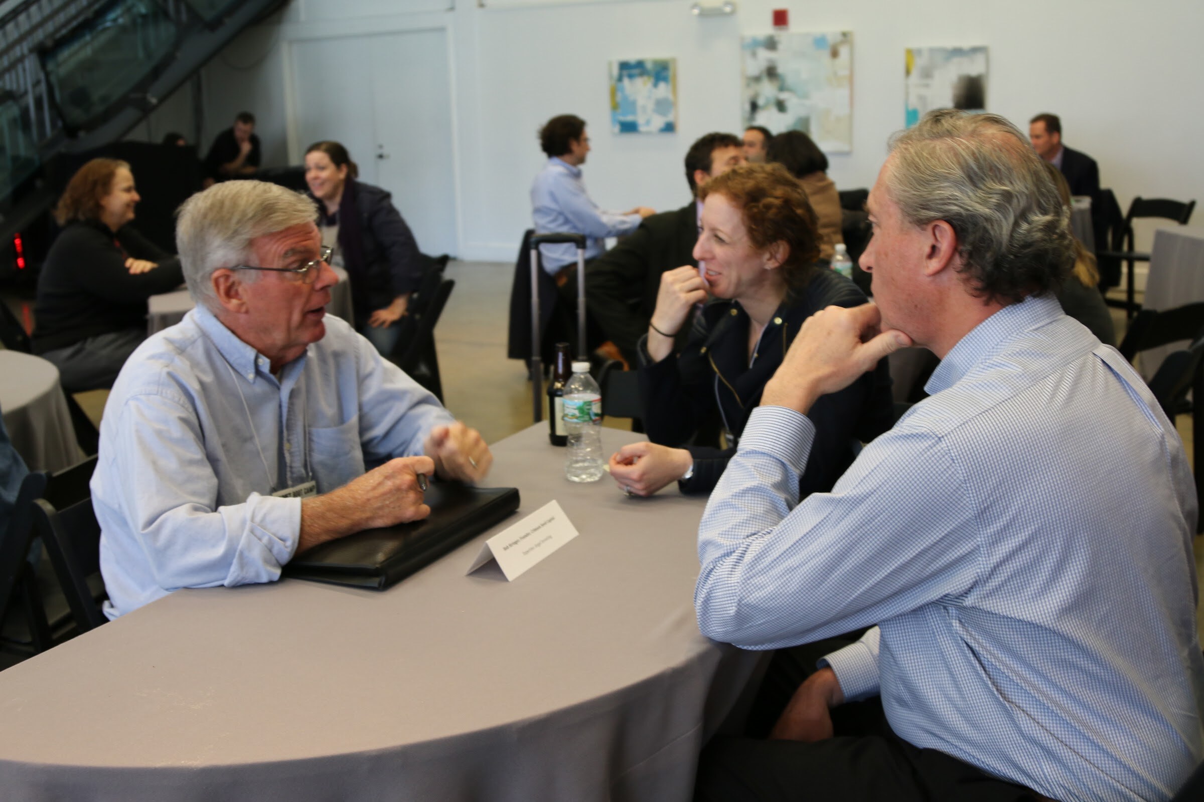 Three attendees in conversation at a table with a name placard, with other meetings going on behind them.