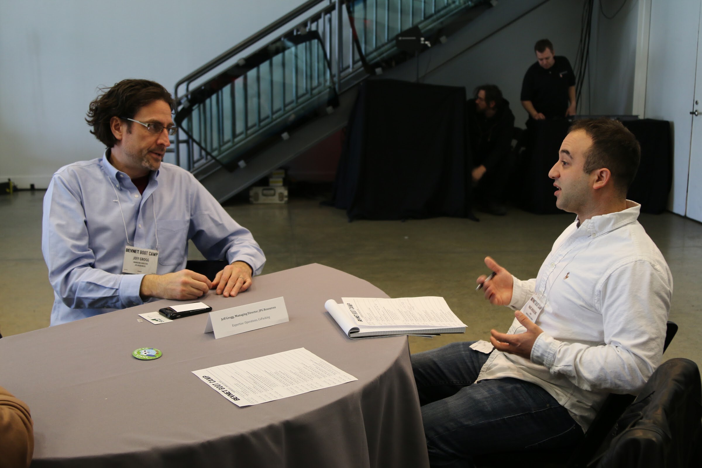 Two men meeting at a table with a name placard and agenda sheets, with event staff near the staircase behind them.