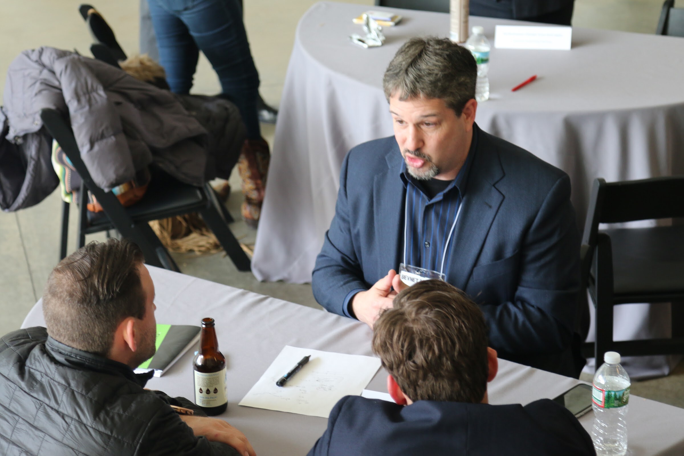 Overhead view of man in navy blazer speaking with two attendees at table with beer bottle.