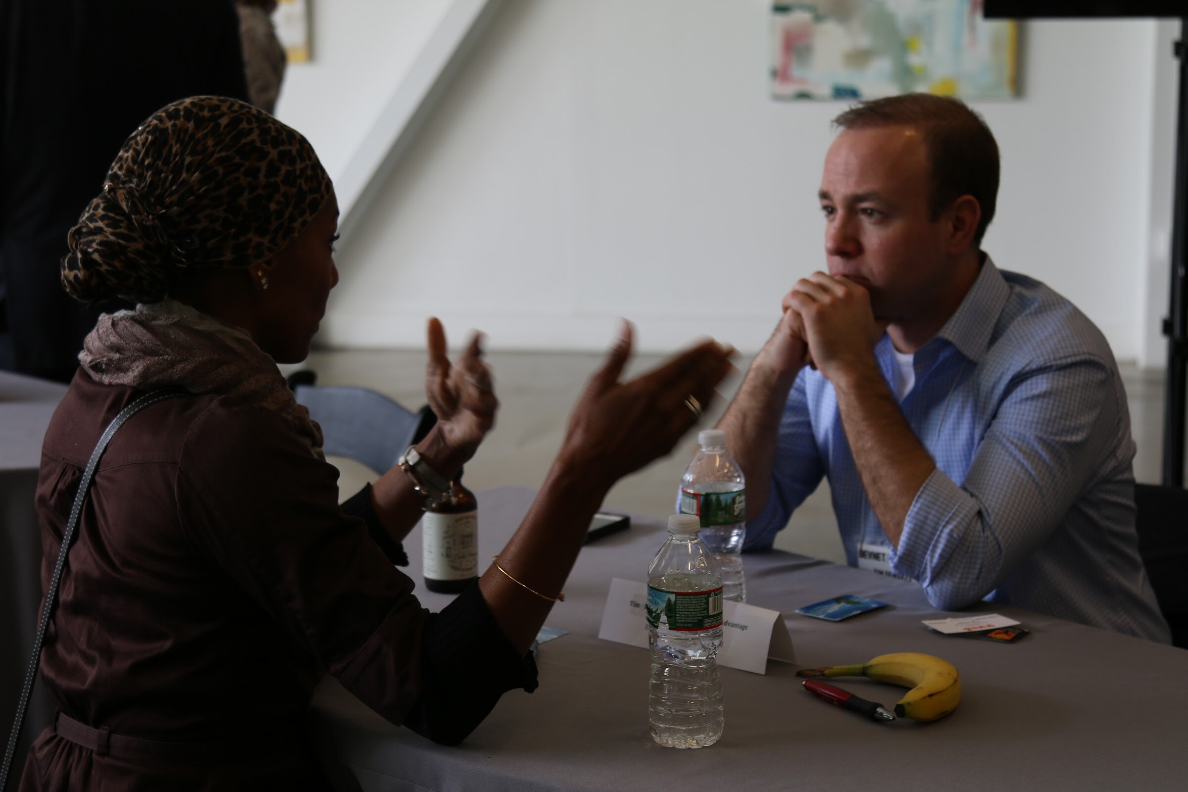 Woman gestures while man in blue shirt listens, hands on chin, banana and water on table.