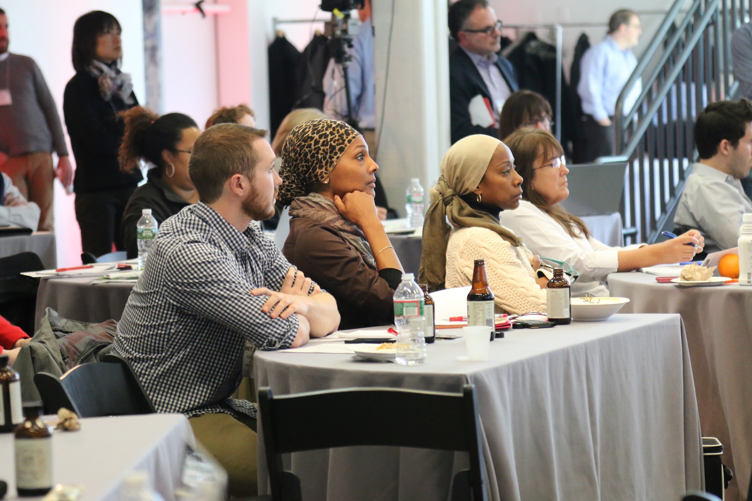 Attendees seated at table listening, one woman in leopard head wrap, hand on chin.