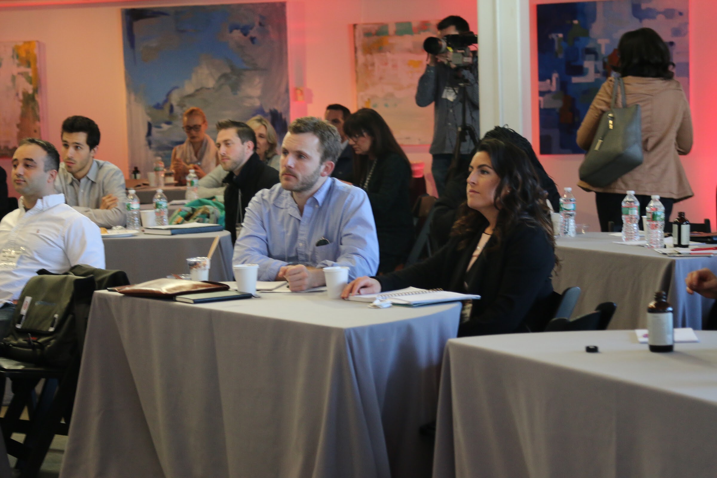 Attendees seated at tables watching a presentation, with a video camera operator behind them.