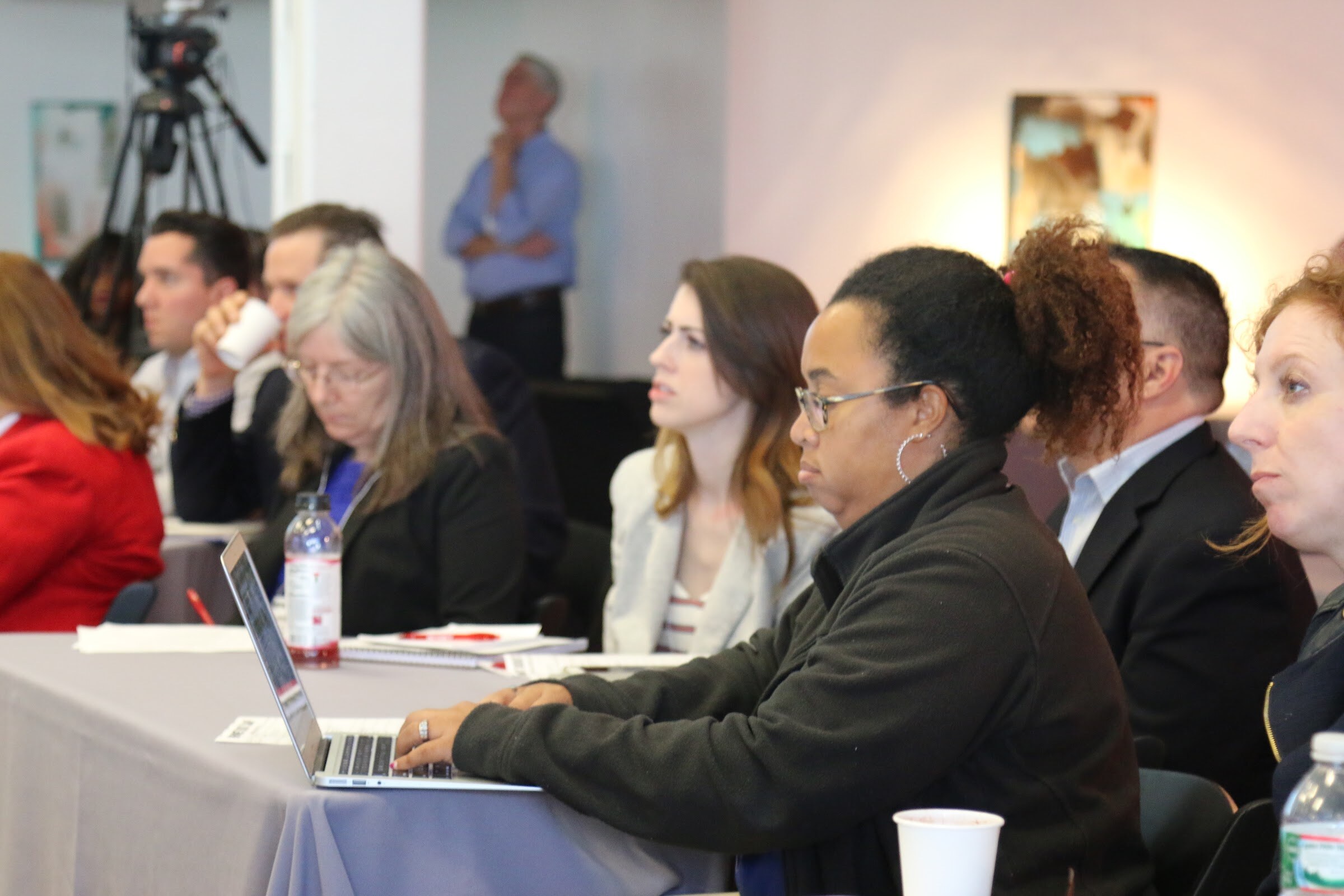 Attendees seated at table, woman typing on laptop, others watching presentation.