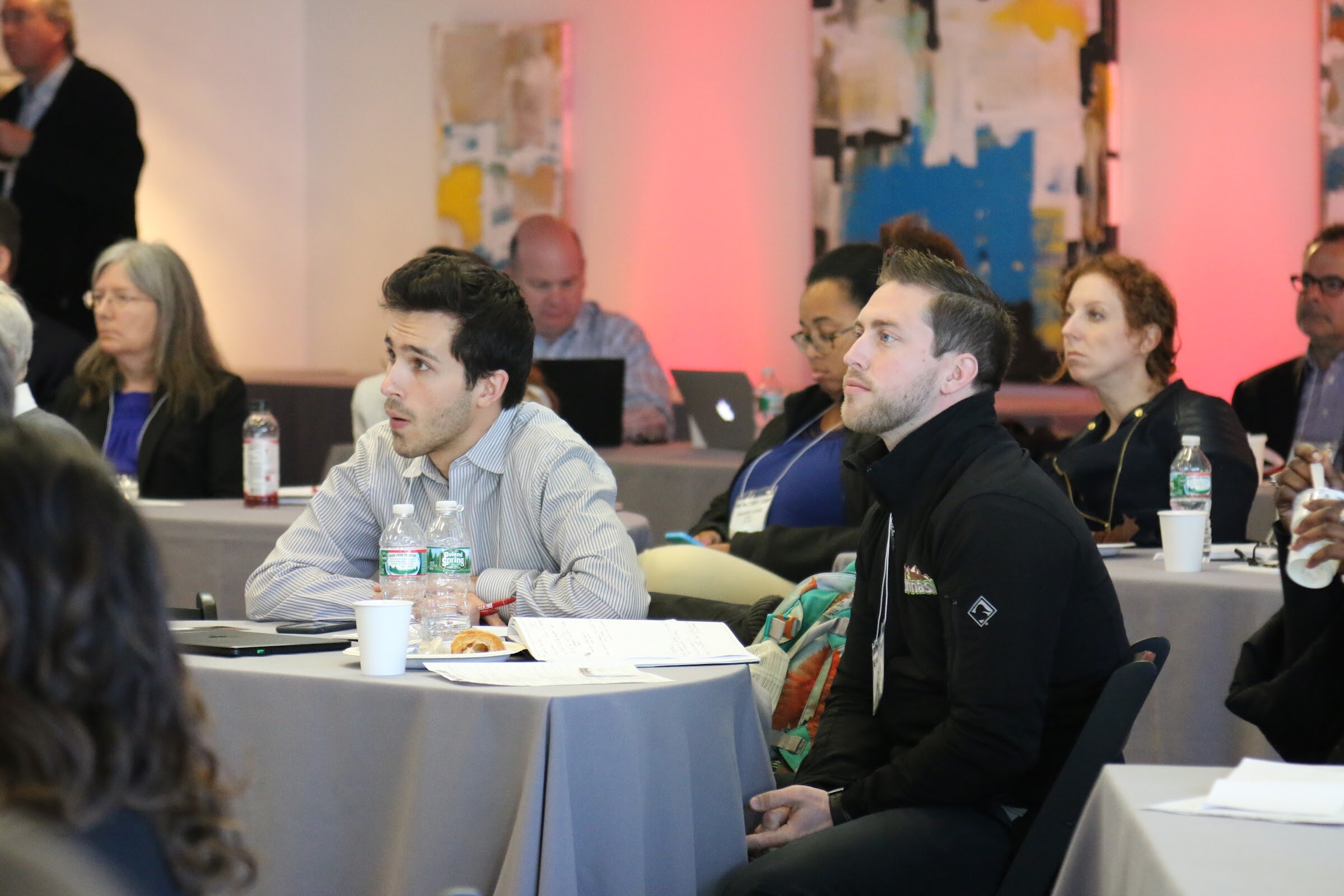 Attendees seated listening, two men front, pink lit art walls.