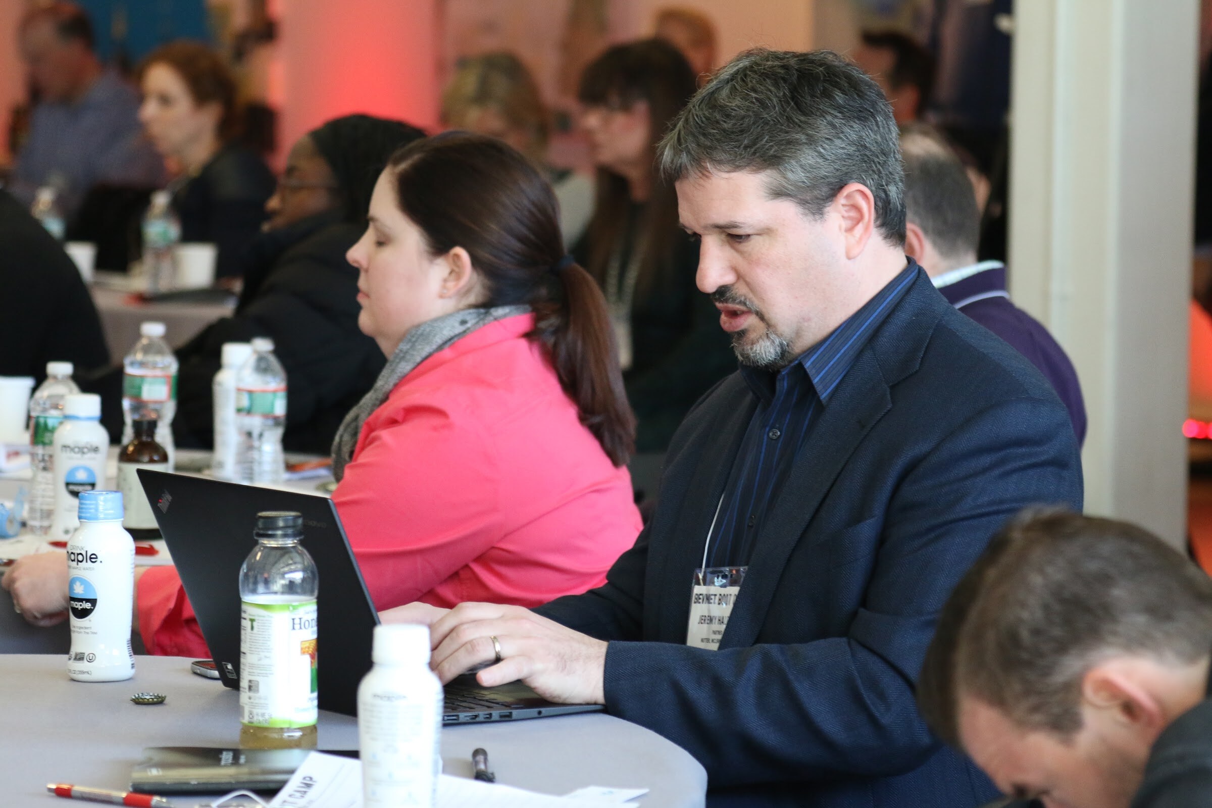 Attendee typing on a laptop at a table beside a woman in a pink jacket, with Maple bottles in front of them.