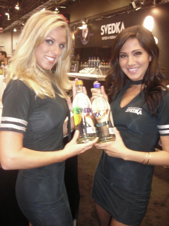 Two smiling women in black Svedka dresses hold up two Svedka vodka bottles in front of a dark Svedka booth with the brand name lit on the wall and bottles on shelves behind them.