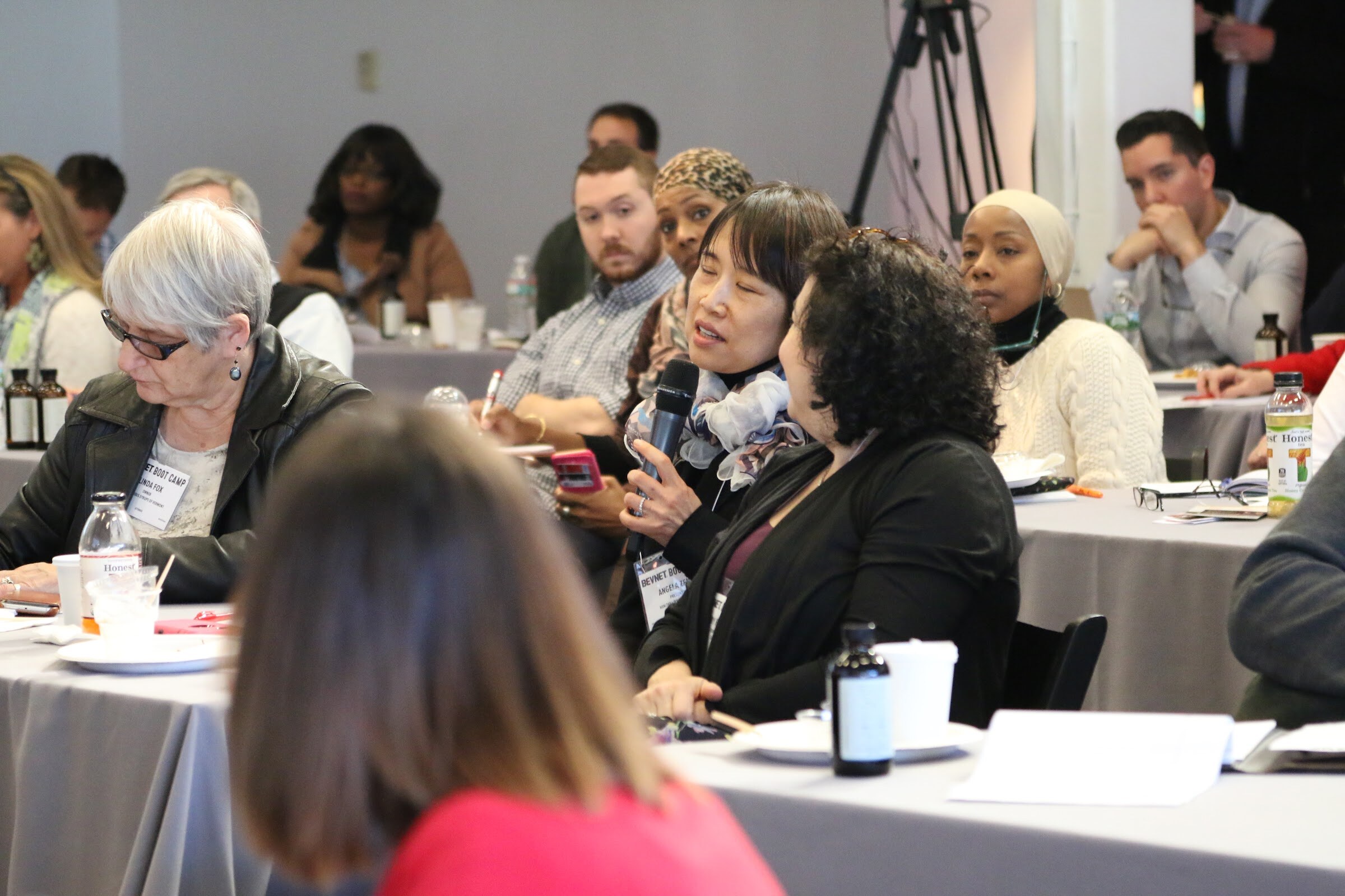 Audience member holding microphone asks question, attendees seated at tables.