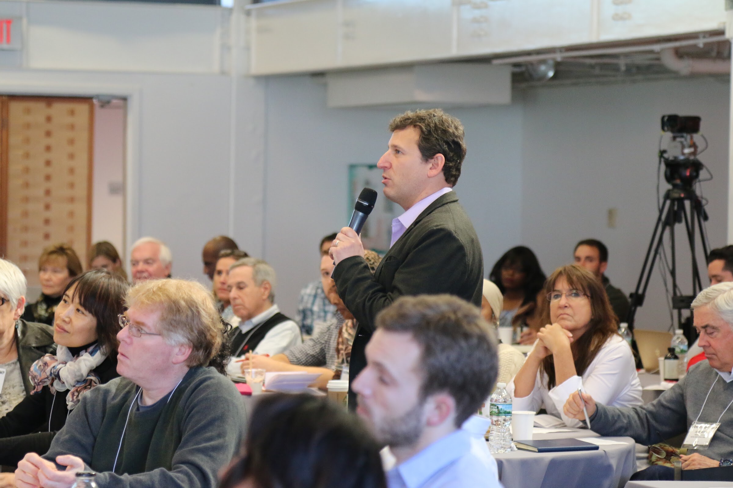 Audience member stands holding microphone asking a question, camera on tripod right.