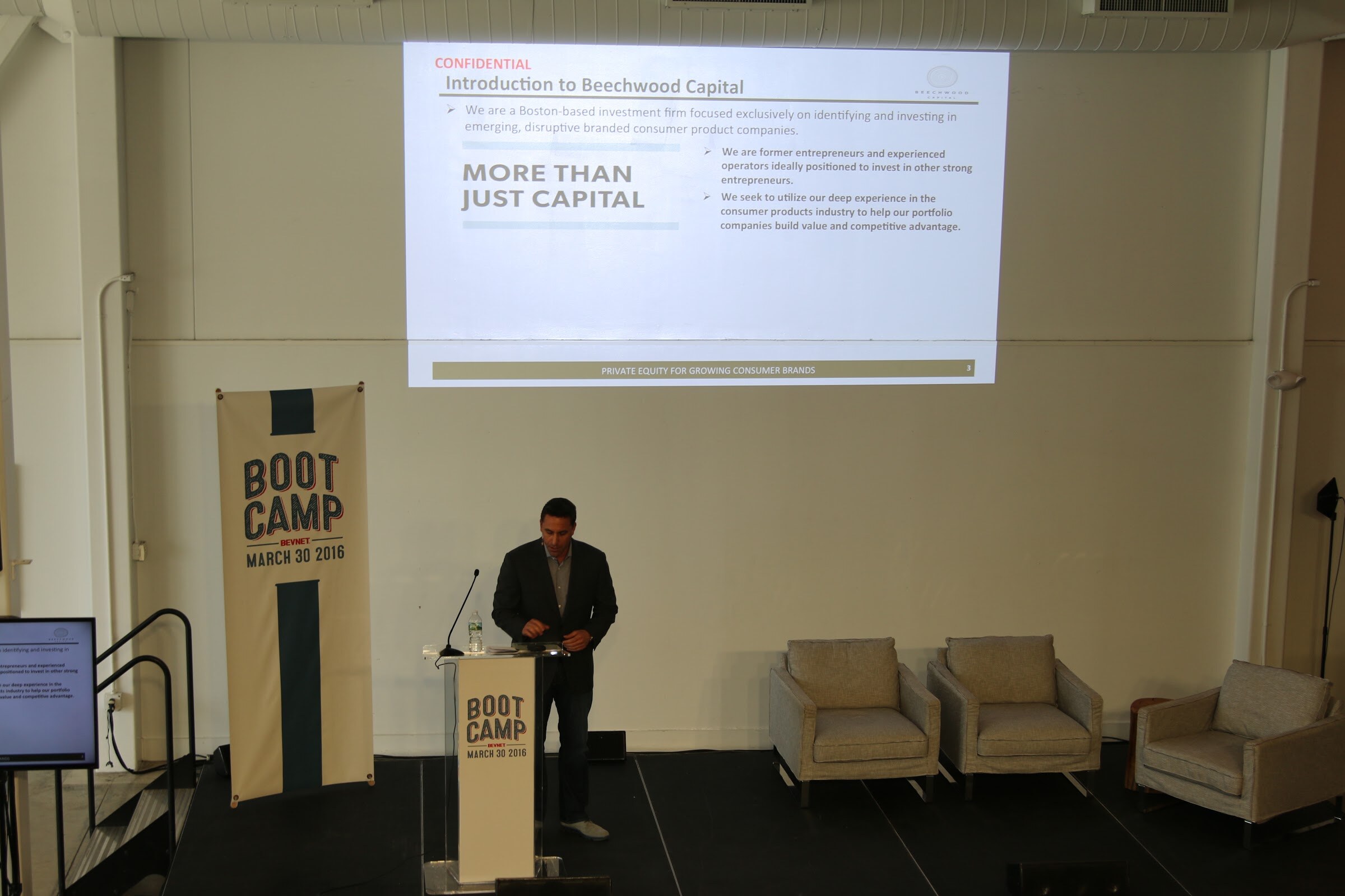 Elevated: presenter at podium, slide Introduction to Beechwood Capital More Than Just Capital, banner and empty armchairs.