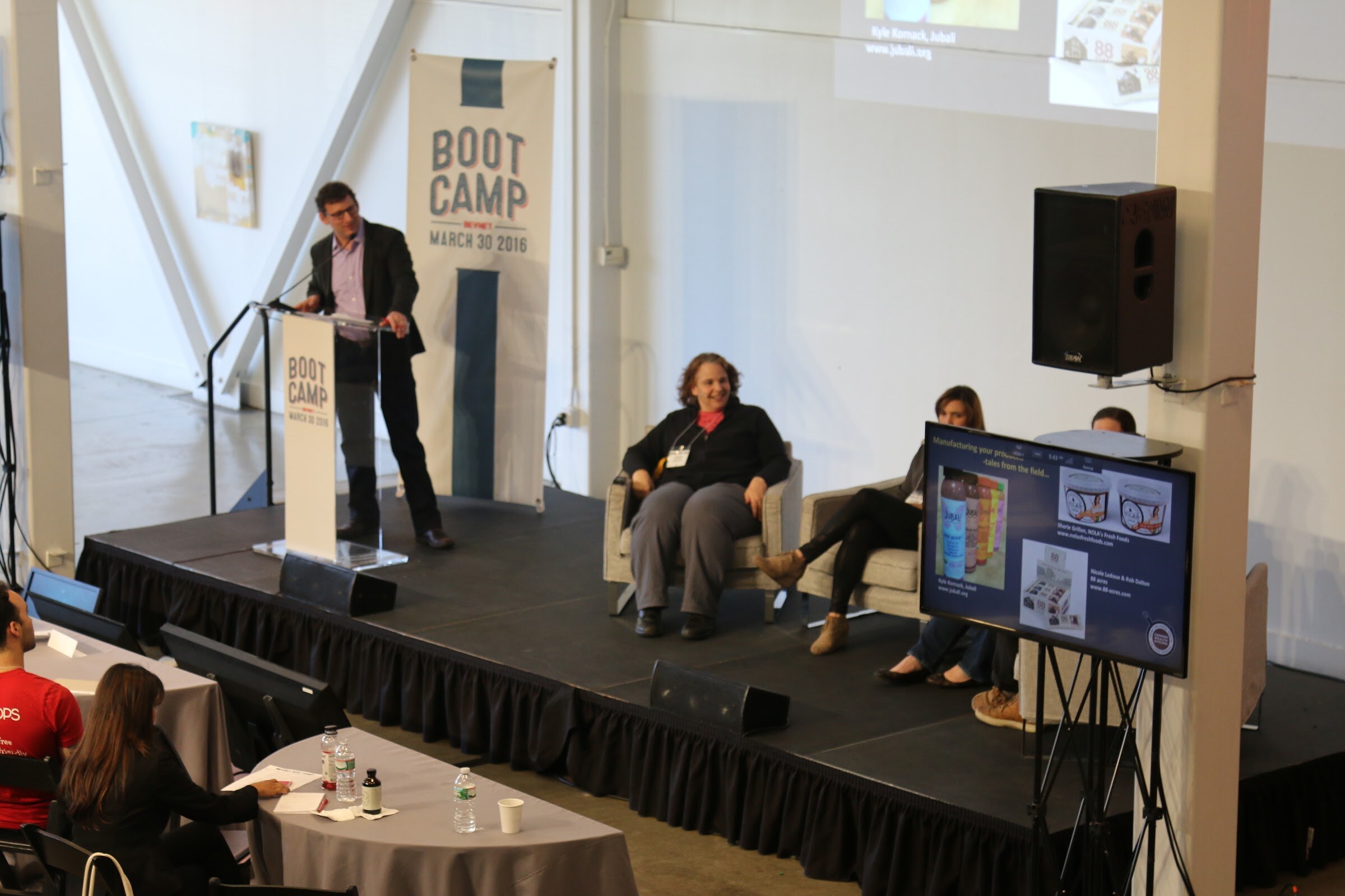 Elevated: speaker at podium, three seated panelists, monitor showing product slide.