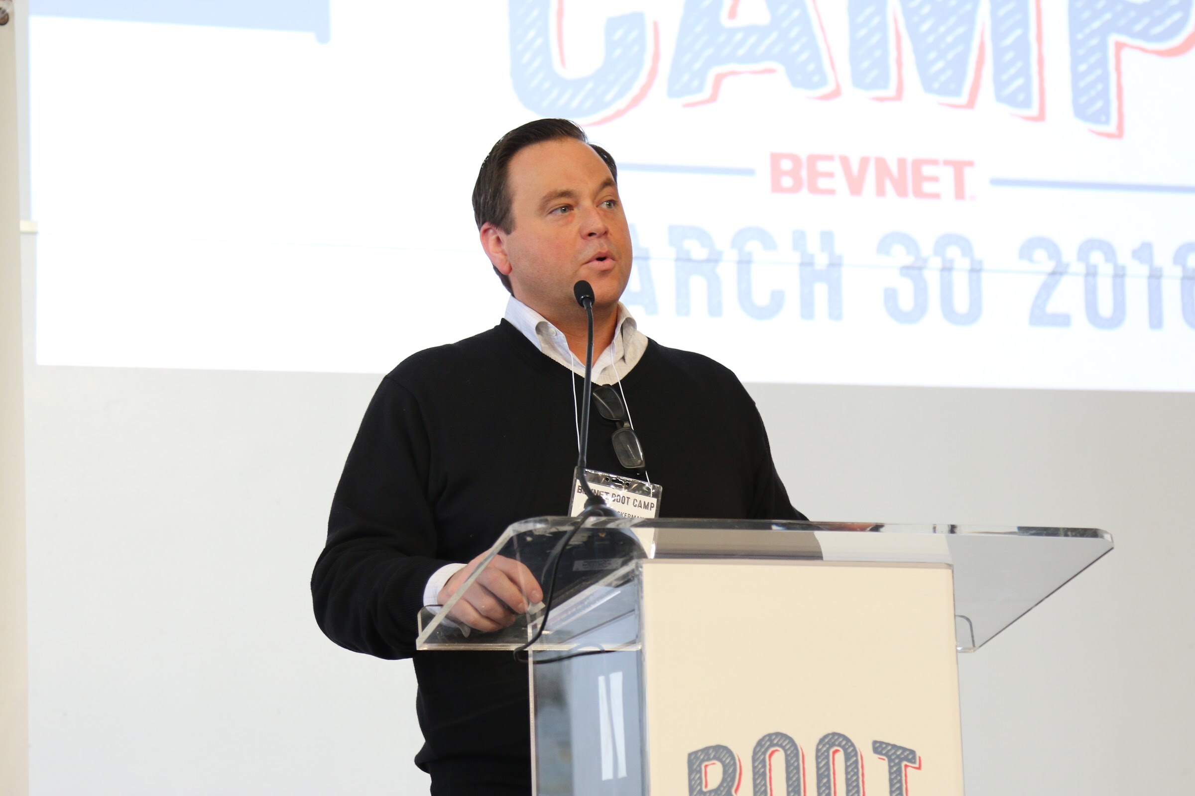 Man in black sweater speaks at podium, Boot Camp BEVNET March 30 2016 slide behind.
