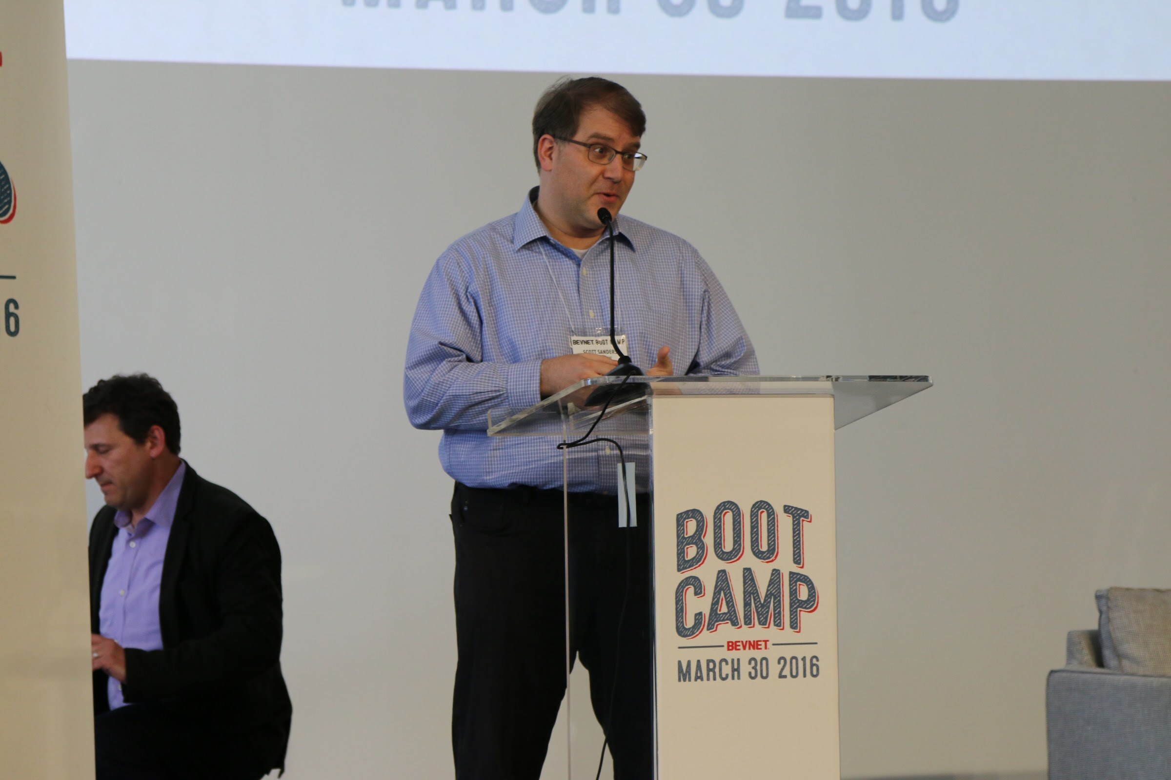 Man in blue shirt speaks at Boot Camp podium, another seated at left.