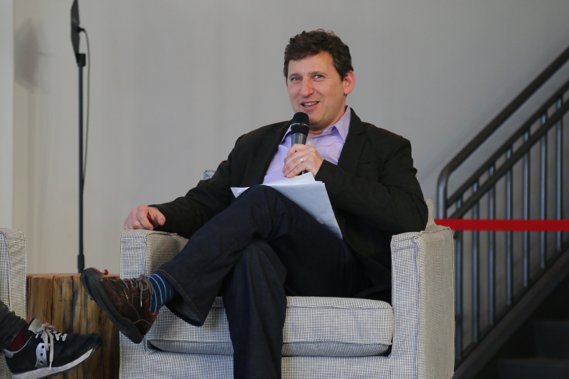 Man seated in an armchair holding a handheld microphone and smiling.