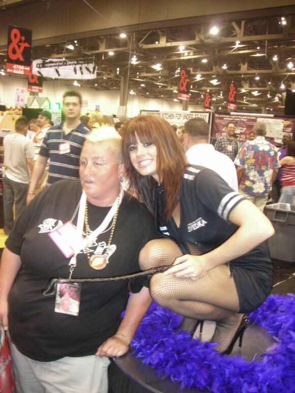 A woman in a black Svedka dress and fishnets crouches on a platform trimmed with purple feathers beside a smiling attendee with a badge and gold necklace, with the show floor behind them.