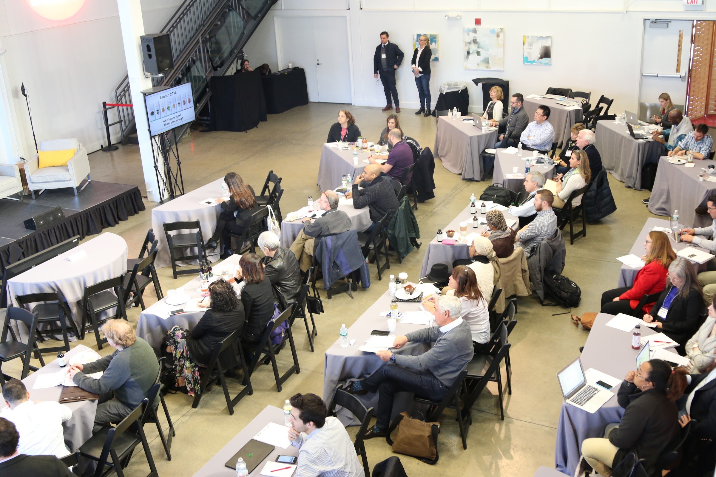 Overhead of room, attendees at tables facing screen showing Launch 2014, two staff standing at back.