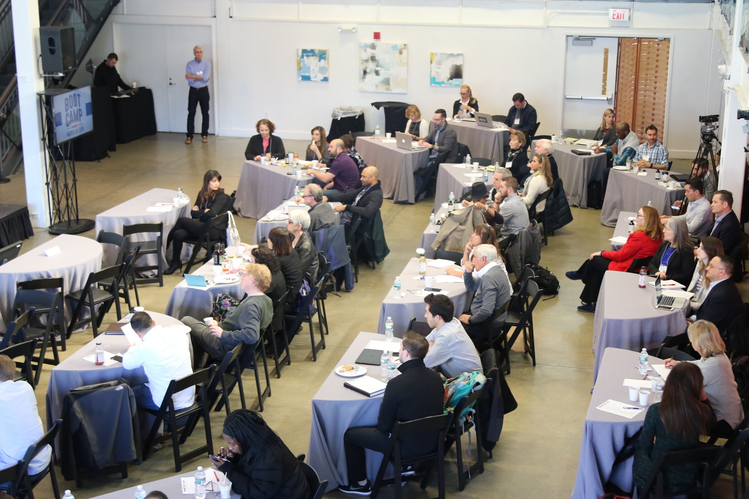 Overhead view of the main room with attendees seated at tables facing the stage and a presenter standing at the back left.