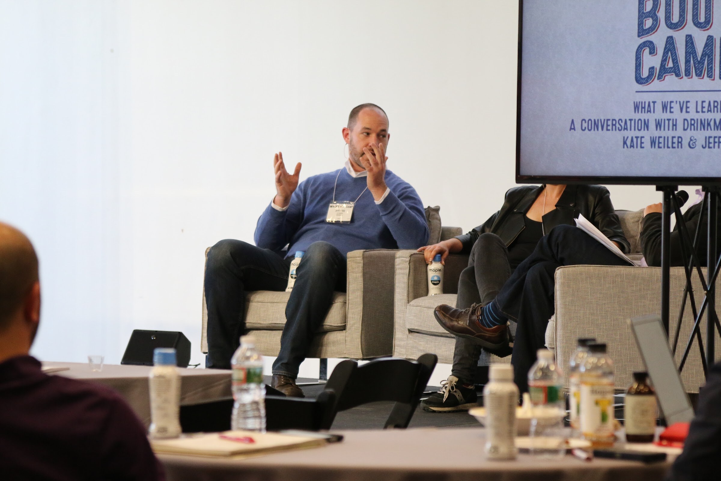 Panelist in blue sweater gesturing, screen shows What We've Learned A Conversation with Drinkmaker..., attendee tables foreground.