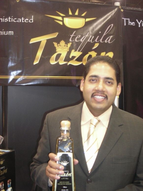 A smiling man in a gray suit and striped tie holds a bottle of Tazón Añejo tequila in front of a black banner with the gold Tazón Tequila logo and crown.