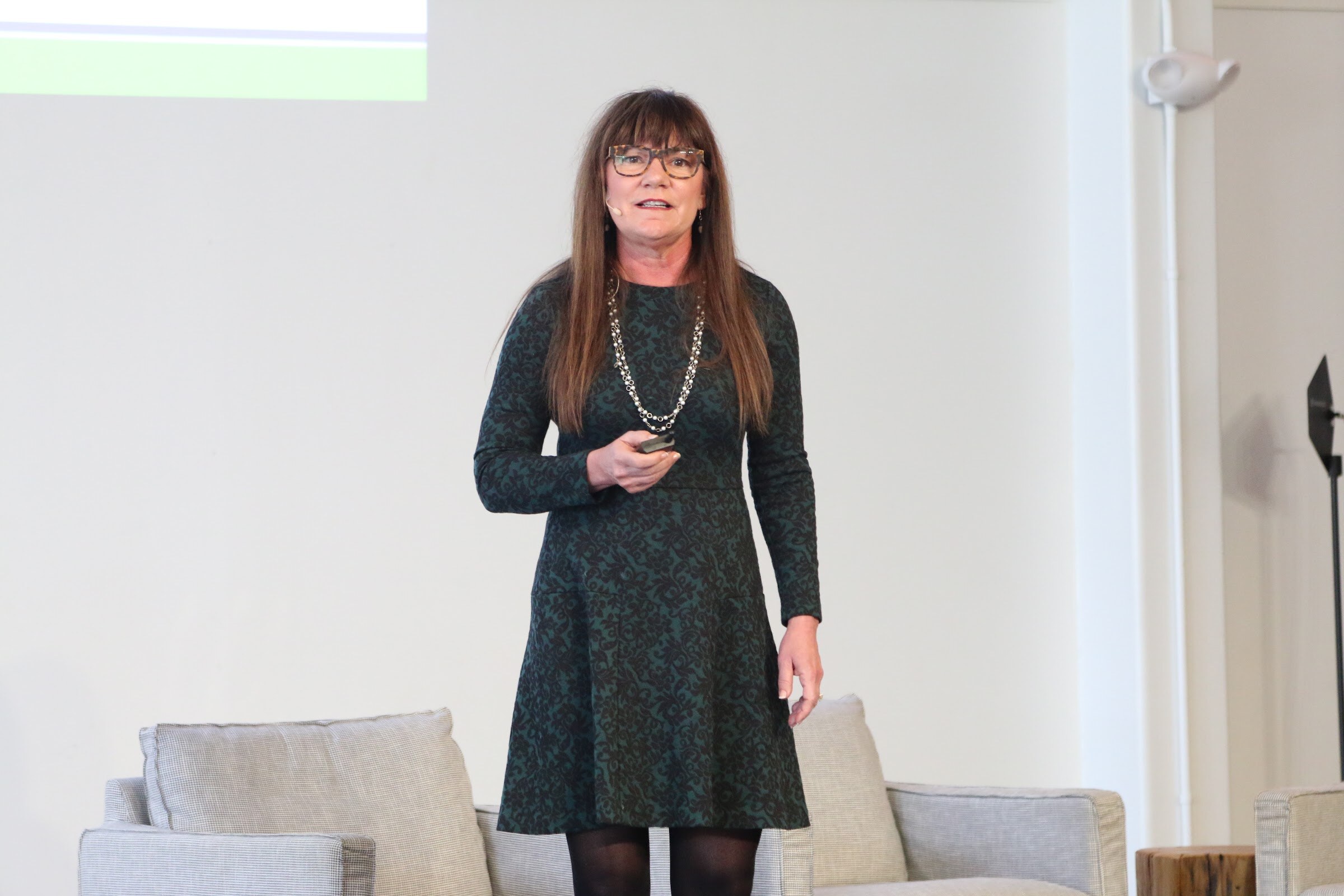 Presenter in dark patterned dress holding clicker, close, speaking.