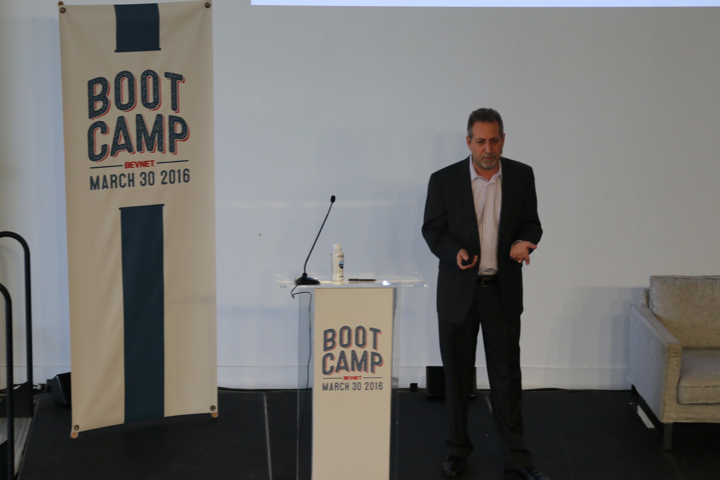 Presenter in dark suit stands beside Boot Camp podium and large banner, gesturing.