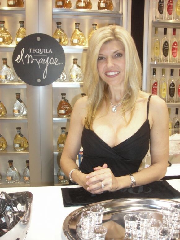 A smiling woman with long blonde hair in a black dress stands behind a white counter with a tray of shot glasses. A Tequila el Mayor sign and shelves of bottles are behind her.