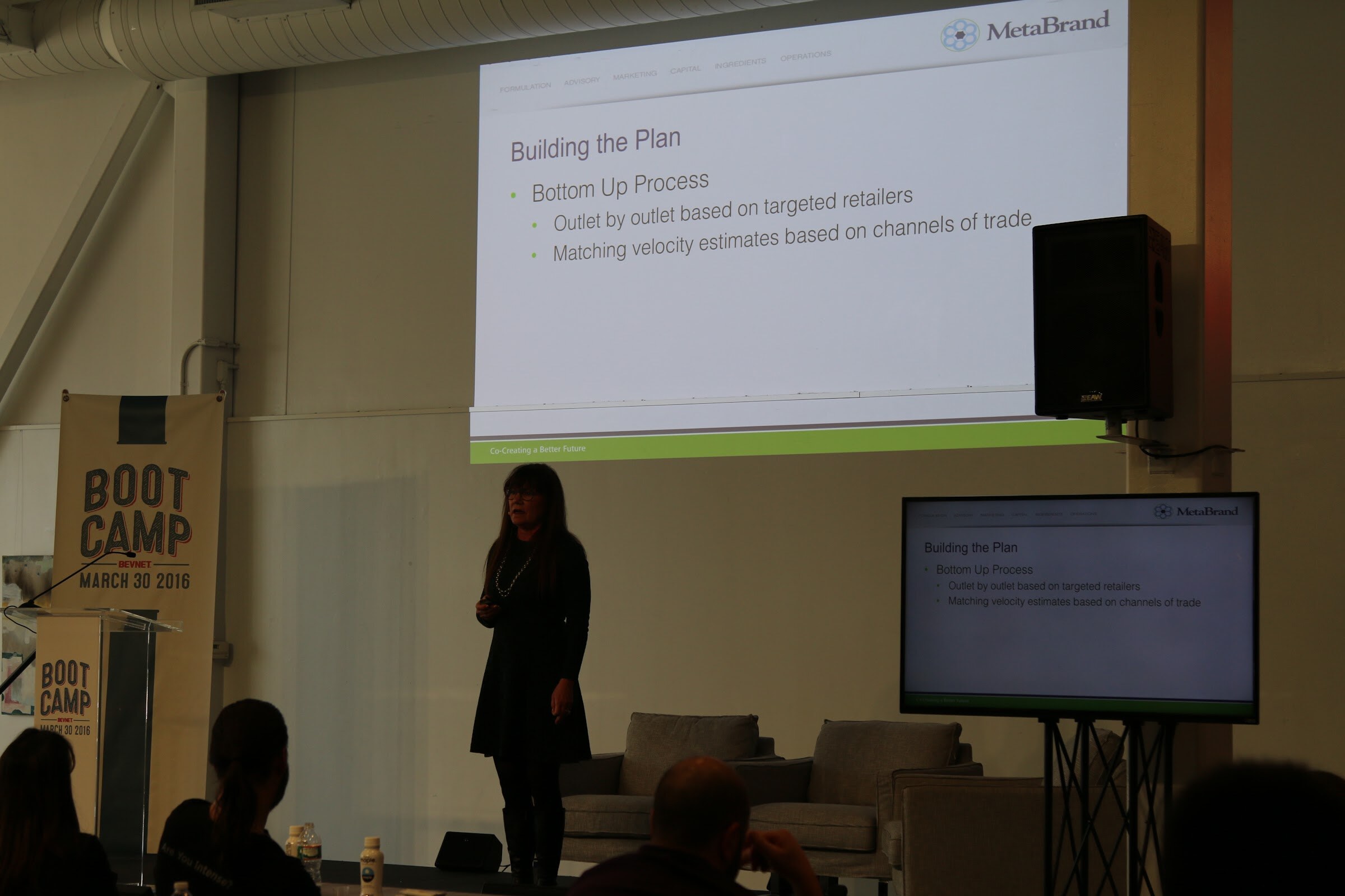 Presenter stands before slide Building the Plan Bottom Up Process (MetaBrand logo), monitor repeats slide.