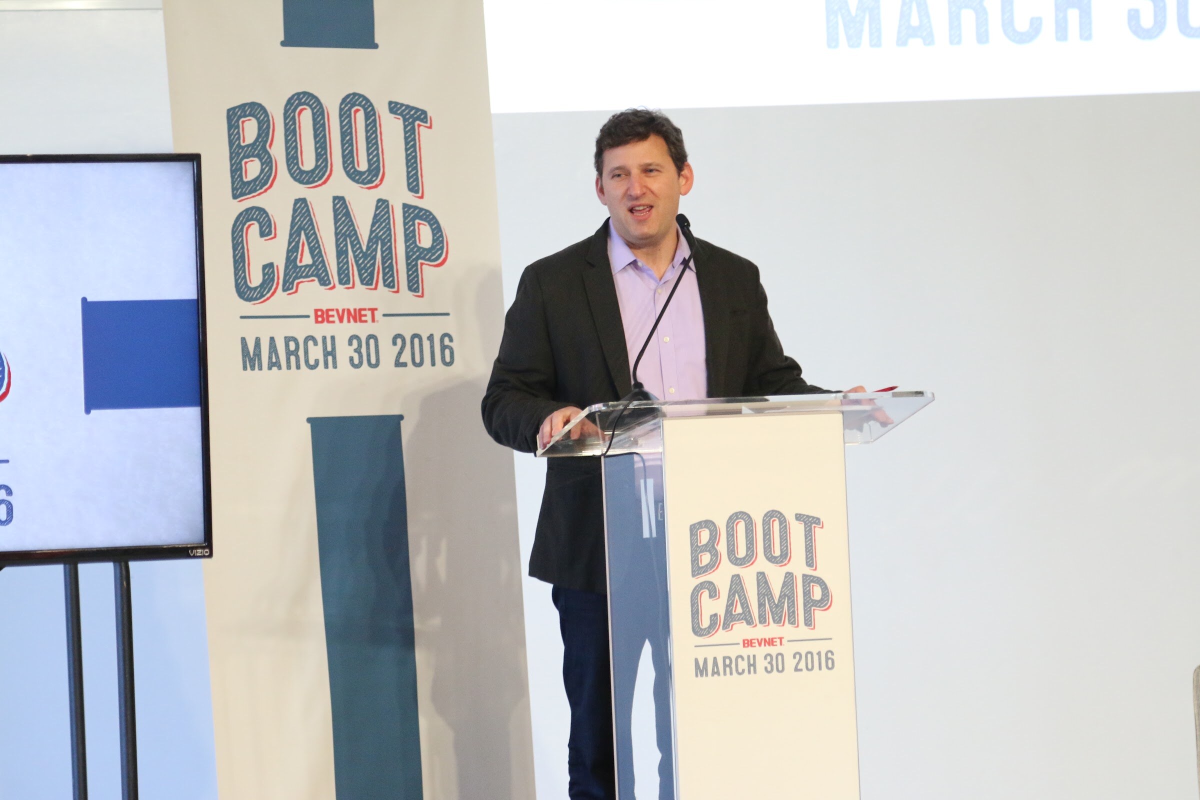 Speaker at podium beside BevNET Boot Camp March 30 2016 banner and screen, smiling mid-talk.