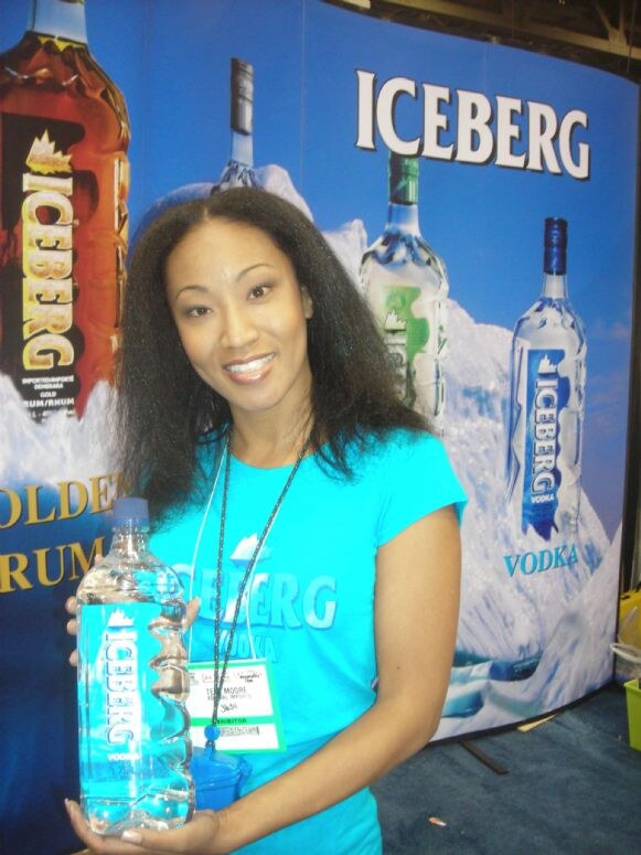 Teri Moore, wearing a teal Iceberg shirt and an exhibitor badge, smiles and holds a bottle of Iceberg Vodka in front of a blue Iceberg backdrop showing rum and vodka bottles.