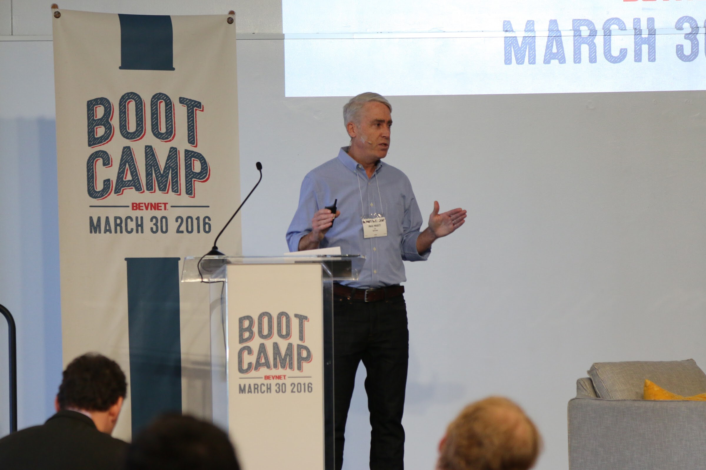 Speaker gesturing at podium with clicker, Boot Camp banner, name badge.