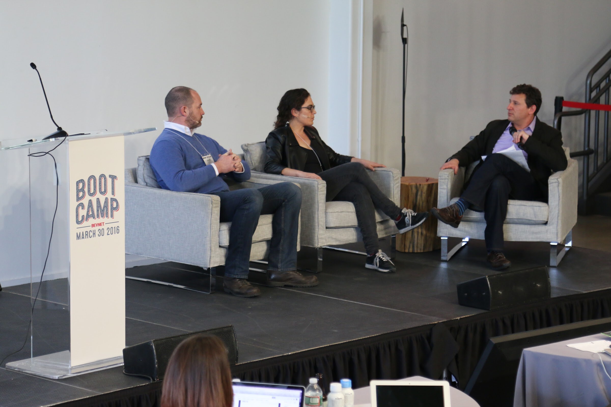 Three people seated on stage in armchairs (two panelists, moderator), Boot Camp podium left.