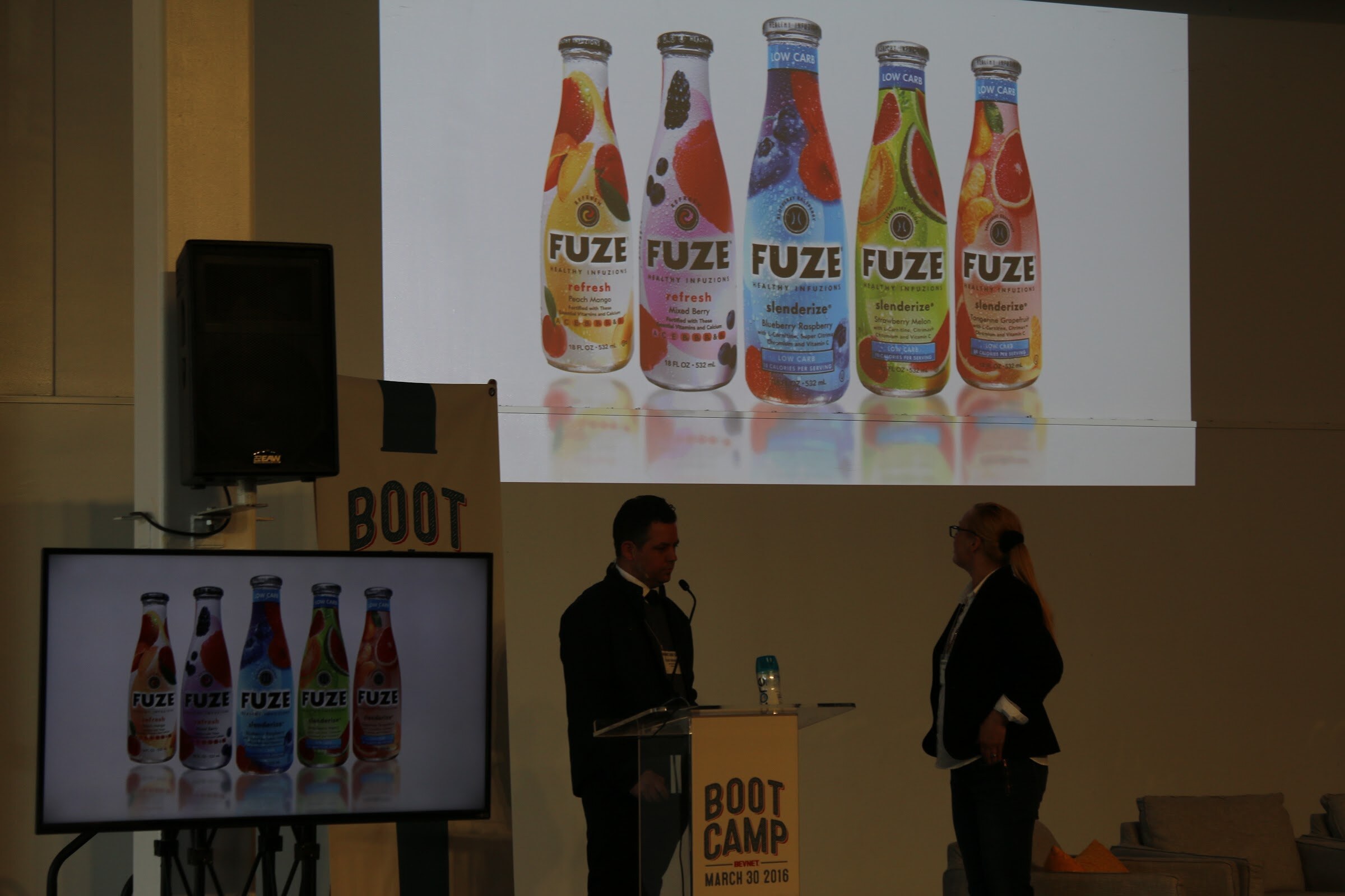 Two presenters beside the podium with a FUZE bottle slide projected on the wall and on the monitor.