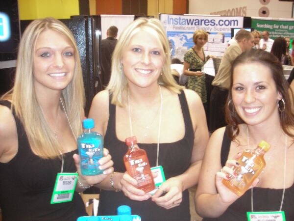 Three smiling women in black tops with show badges each hold a small bottle of Tiki Rita, in blue, red and orange. An Instawares.com banner hangs behind them.