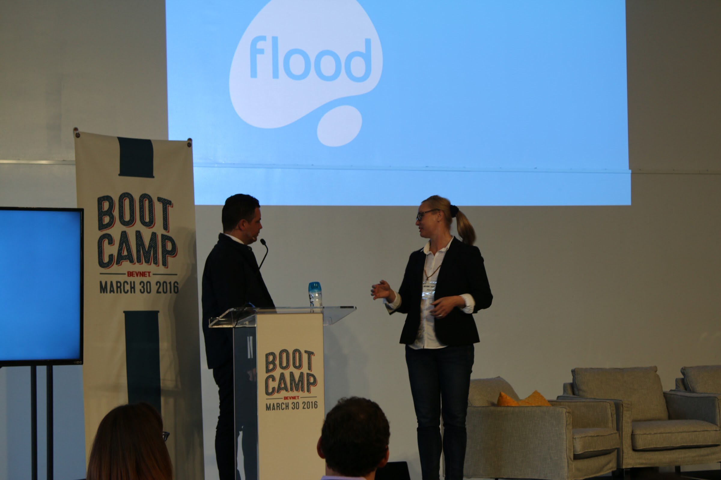 Two presenters on stage beside podium, projected flood logo.
