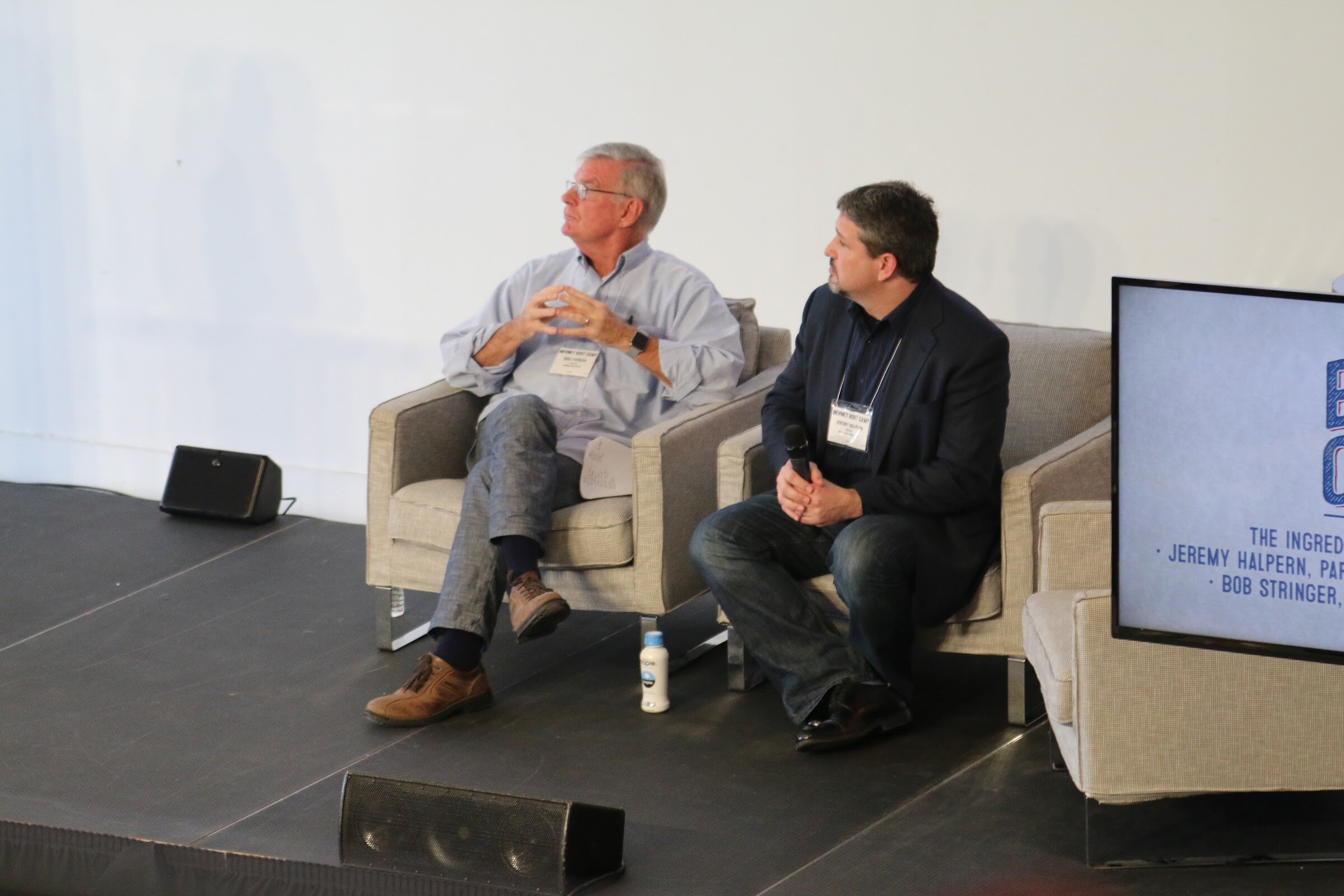 Two seated men on stage, one holding handheld mic, monitor with Ingredients of Angel Investment.