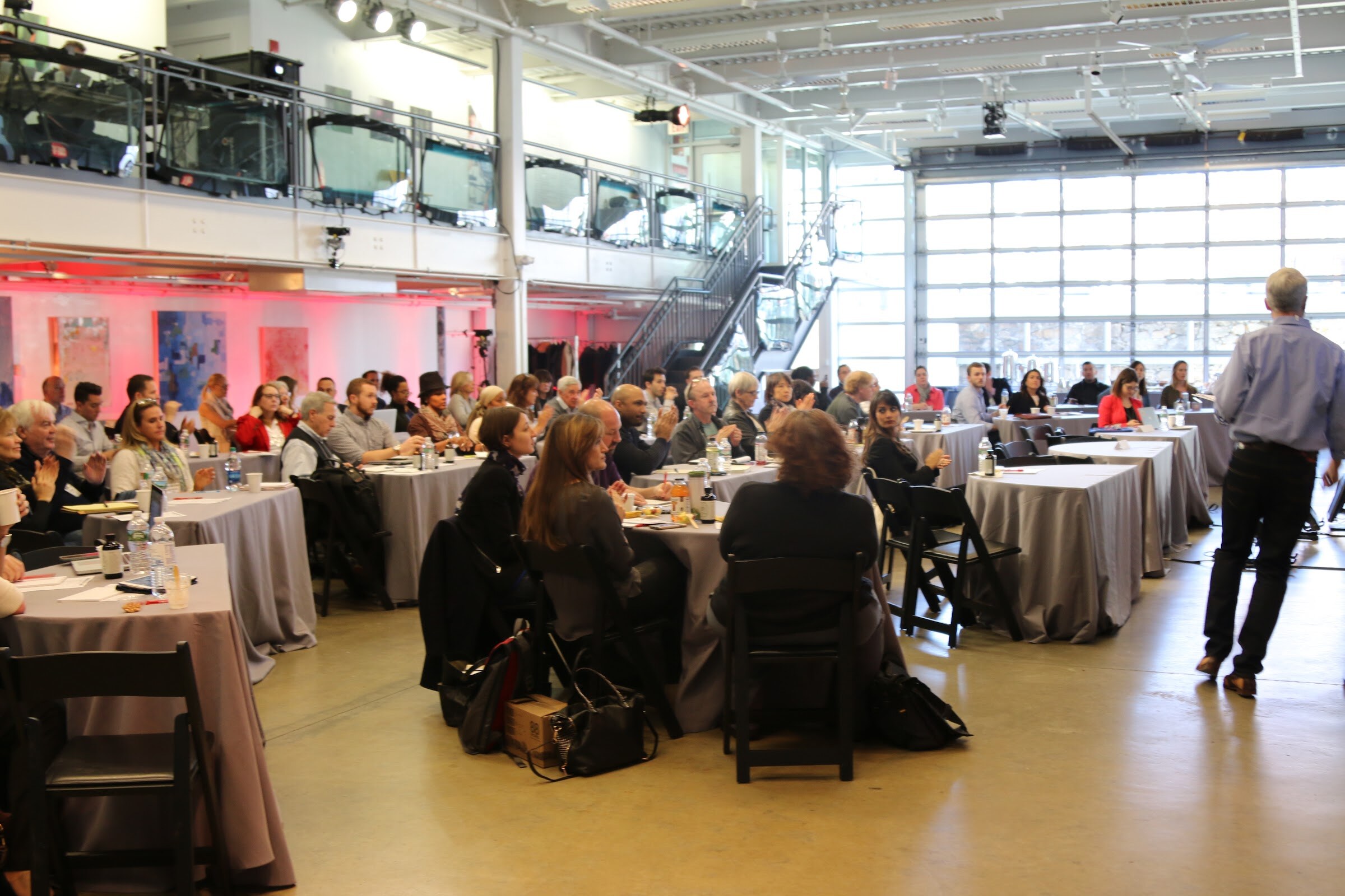 Wide room audience, speaker walking right, balcony and garage doors.