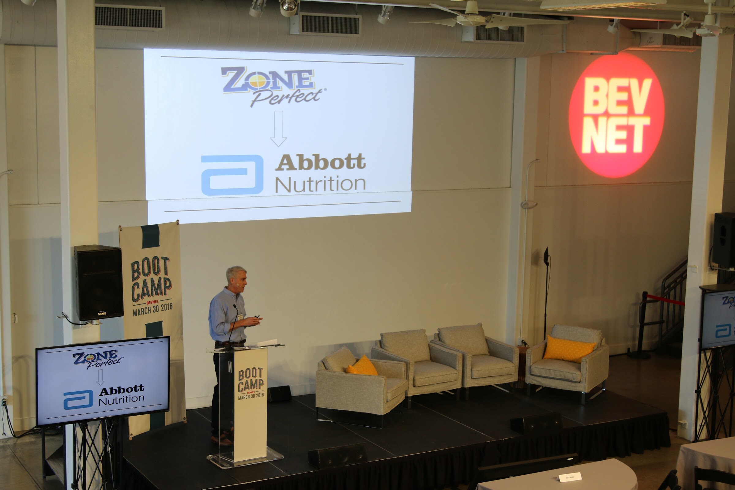 Wide: speaker at podium under projected slide Zone Perfect arrow Abbott Nutrition, BevNET logo on wall, empty armchairs.