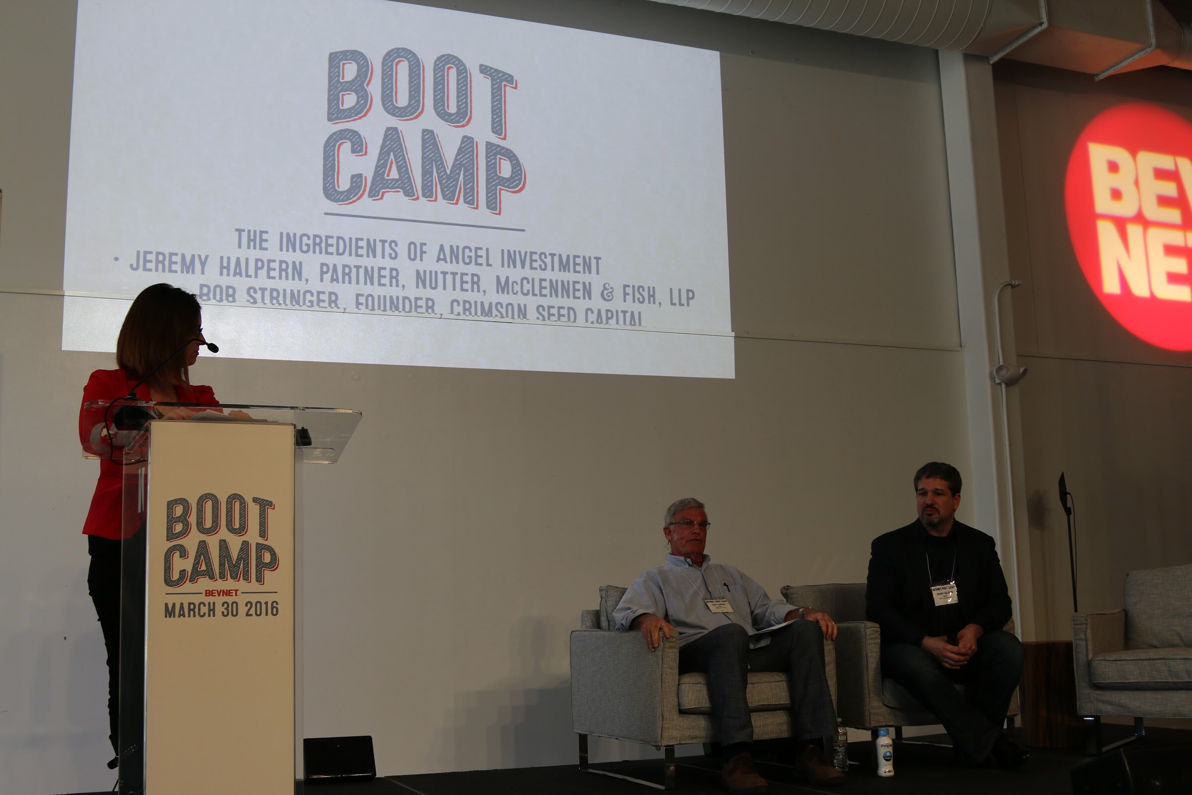 Woman at the podium with two seated speakers, a screen reading The Ingredients of Angel Investment, and the BevNET logo on the wall.