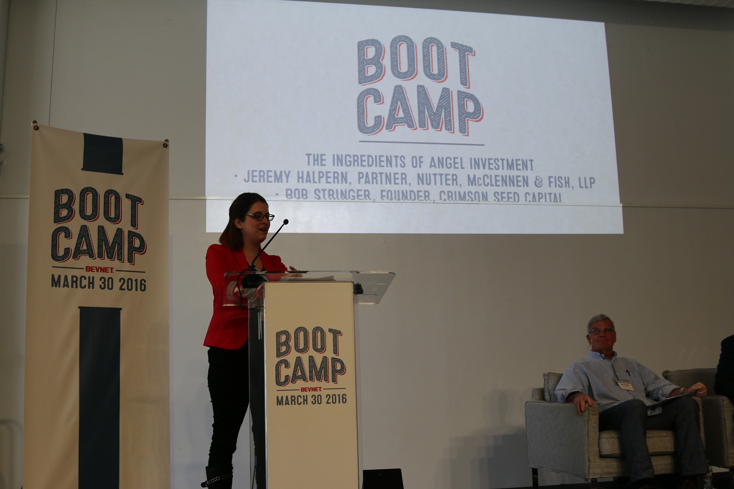Woman in a red blazer speaking at the podium, with a screen reading The Ingredients of Angel Investment and a seated speaker at right.