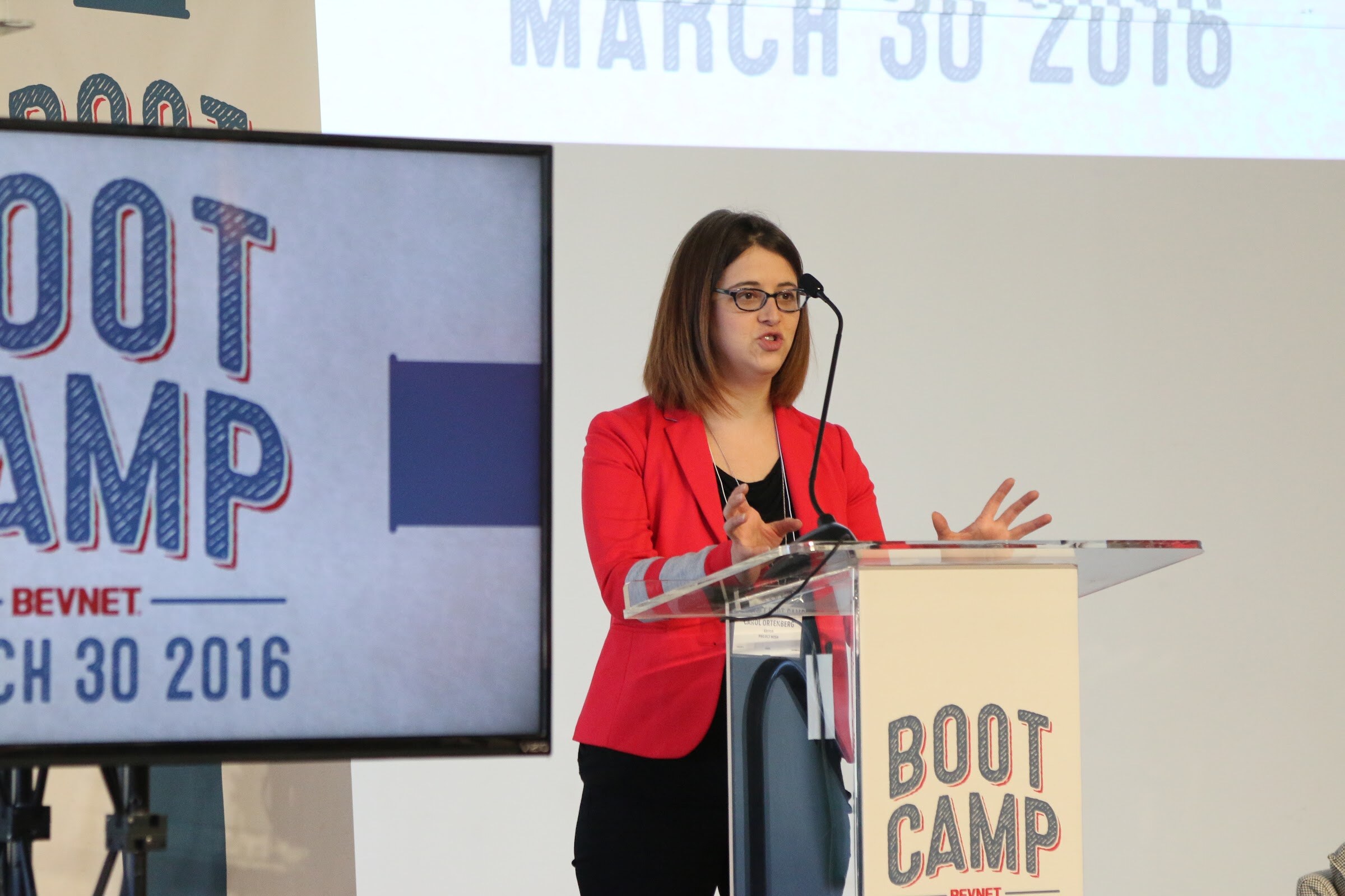 Woman in red blazer gesturing at podium next to Boot Camp screen.