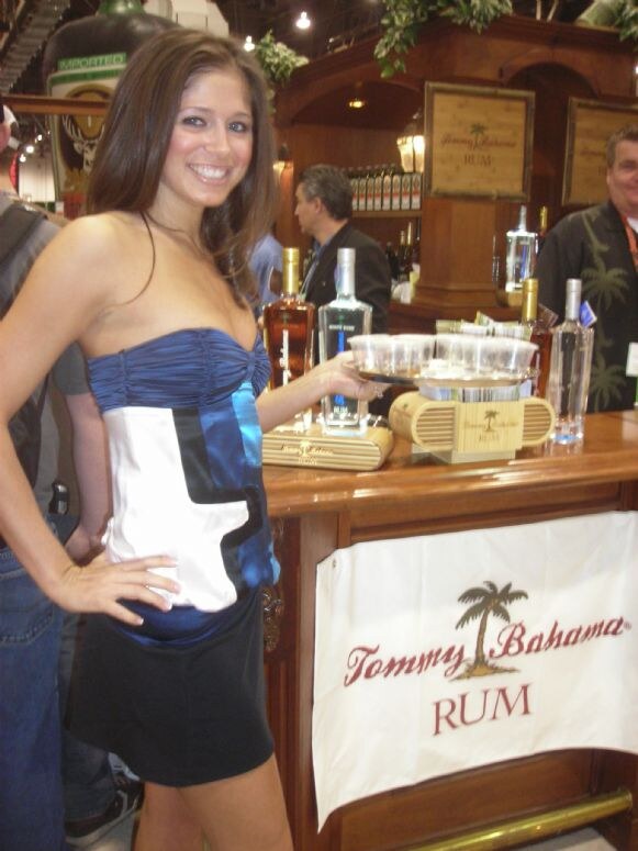A smiling woman in a blue and white strapless dress poses with a hand on her hip beside a wooden bar with bottles, cups and a Tommy Bahama Rum banner.