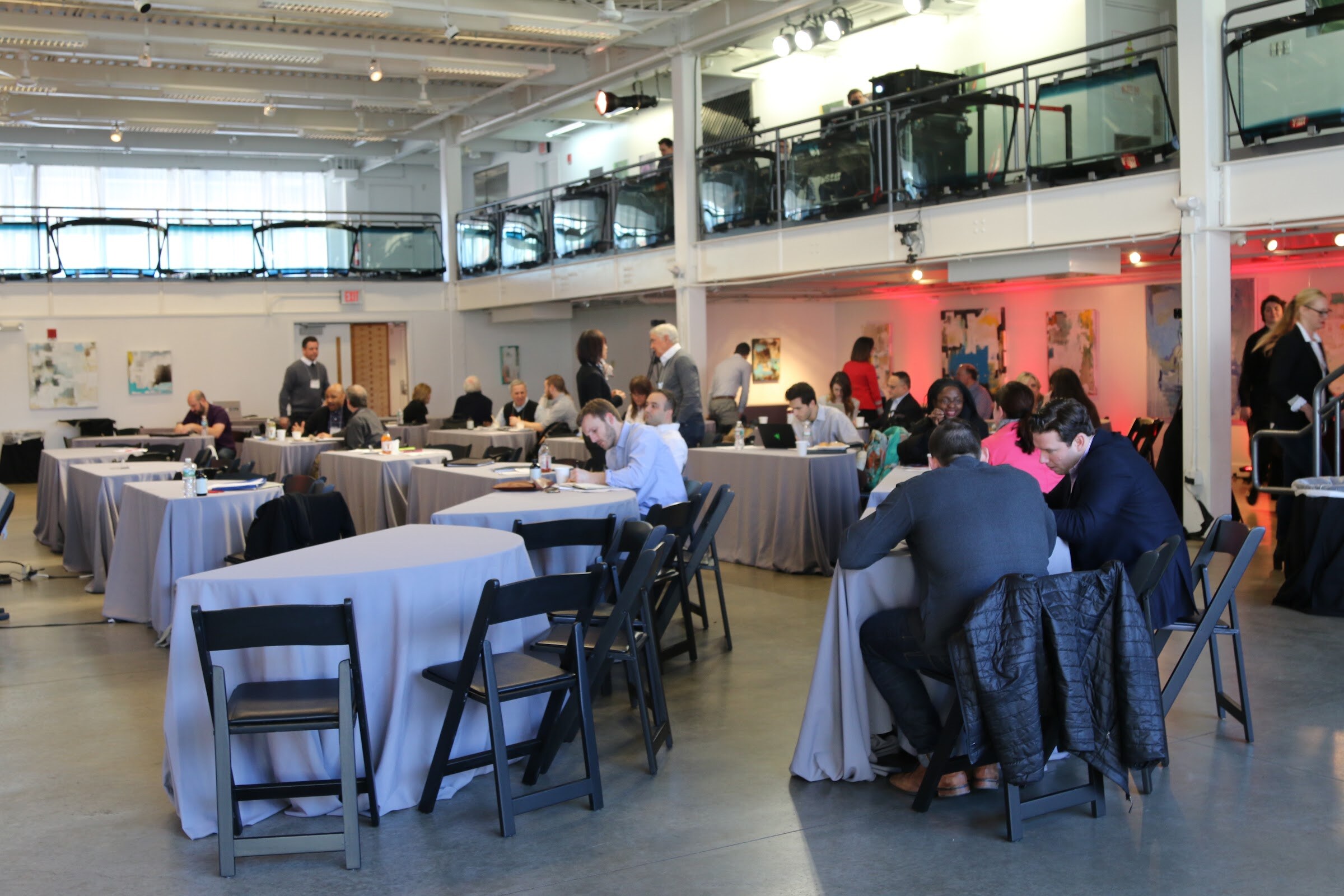 Wide view of the loft venue before sessions: round tables under pink-lit walls, mezzanine with window frames, a few attendees seated.