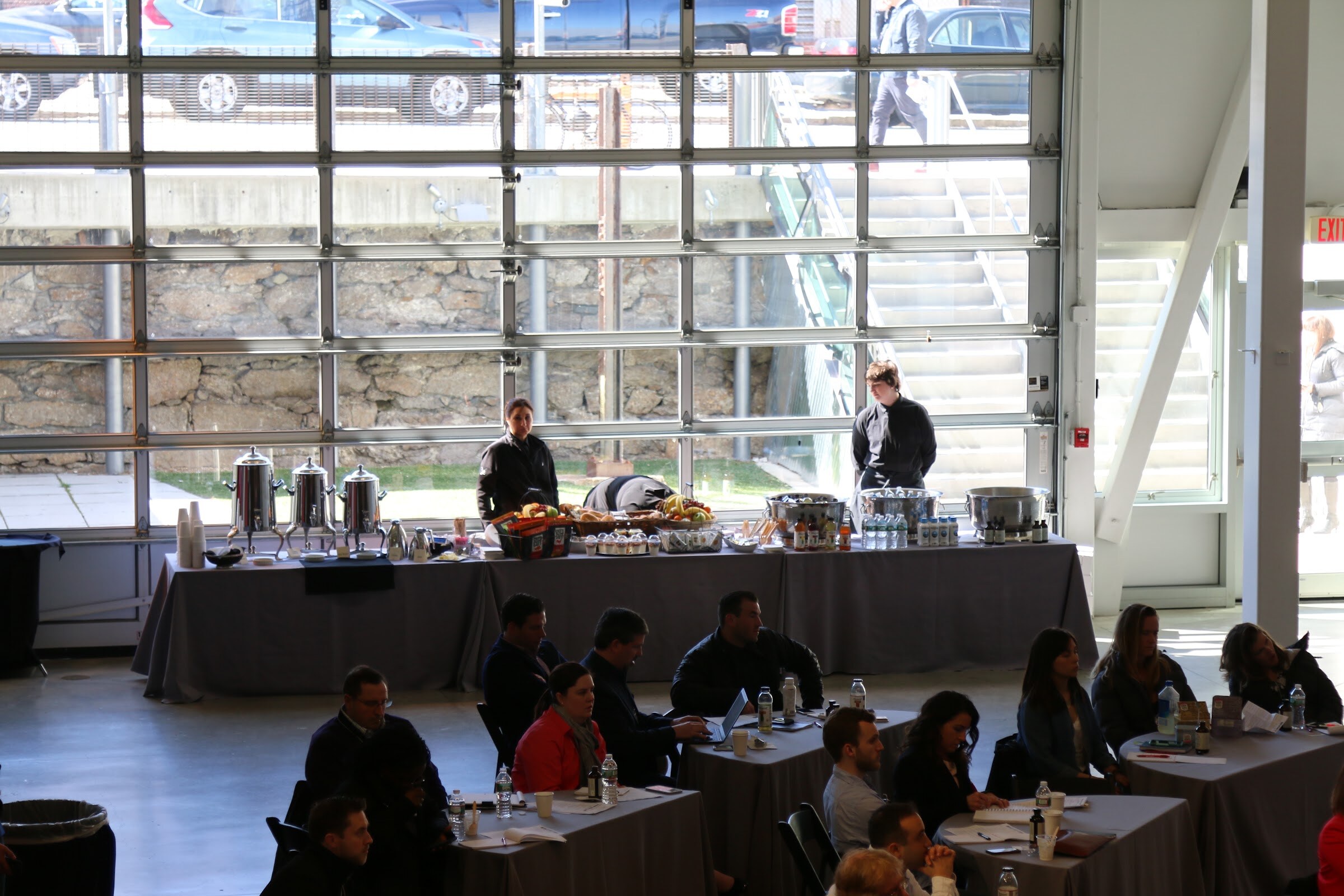 View through large glass garage doors to catering table with coffee urns and fruit, seated attendees below.
