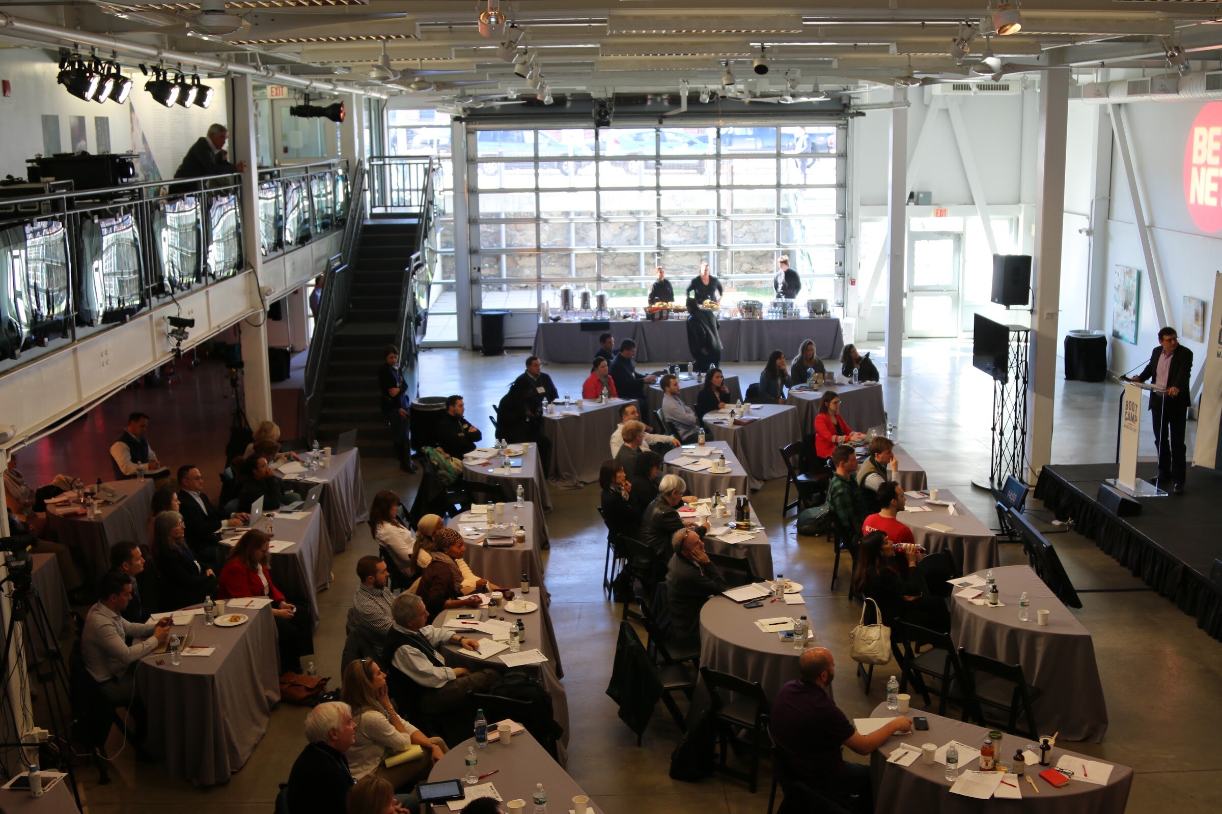 Overhead of room: attendees seated at tables, speaker at podium on right, catering tables back, mezzanine left.