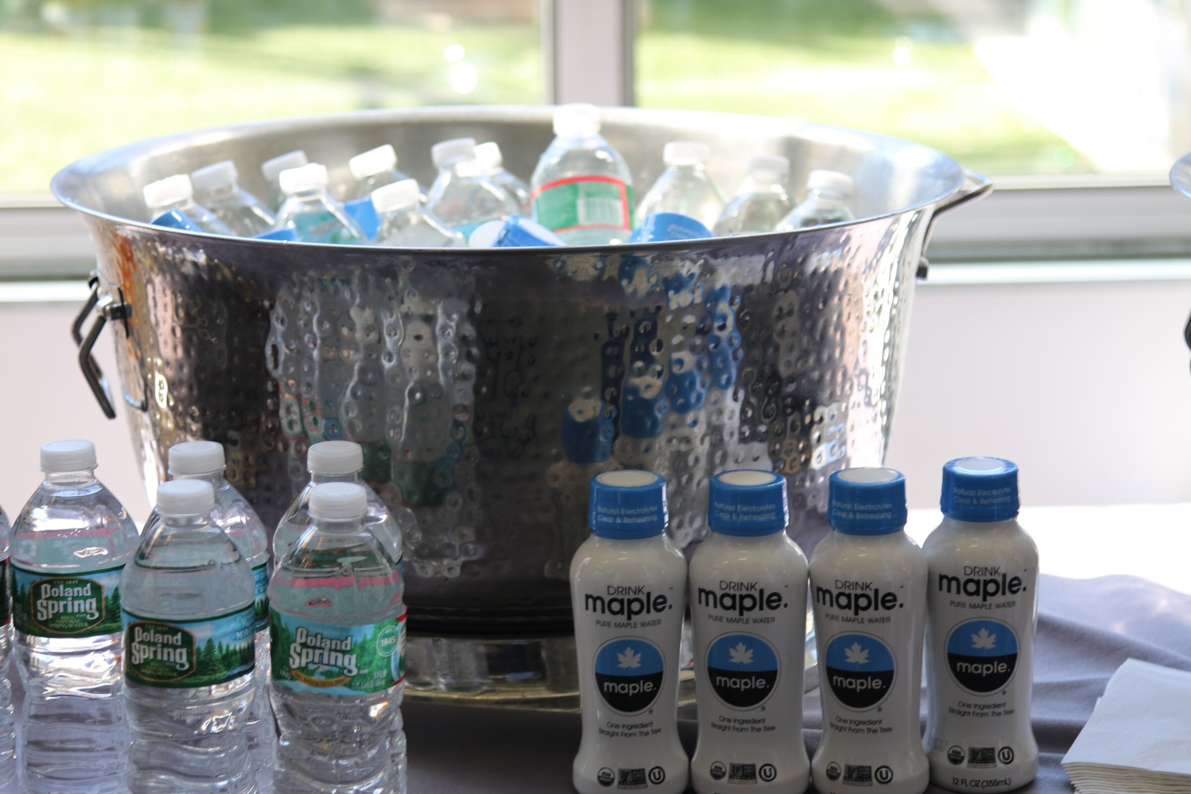 Hammered metal bucket of Poland Spring bottles with four bottles of Drink Maple pure maple water.