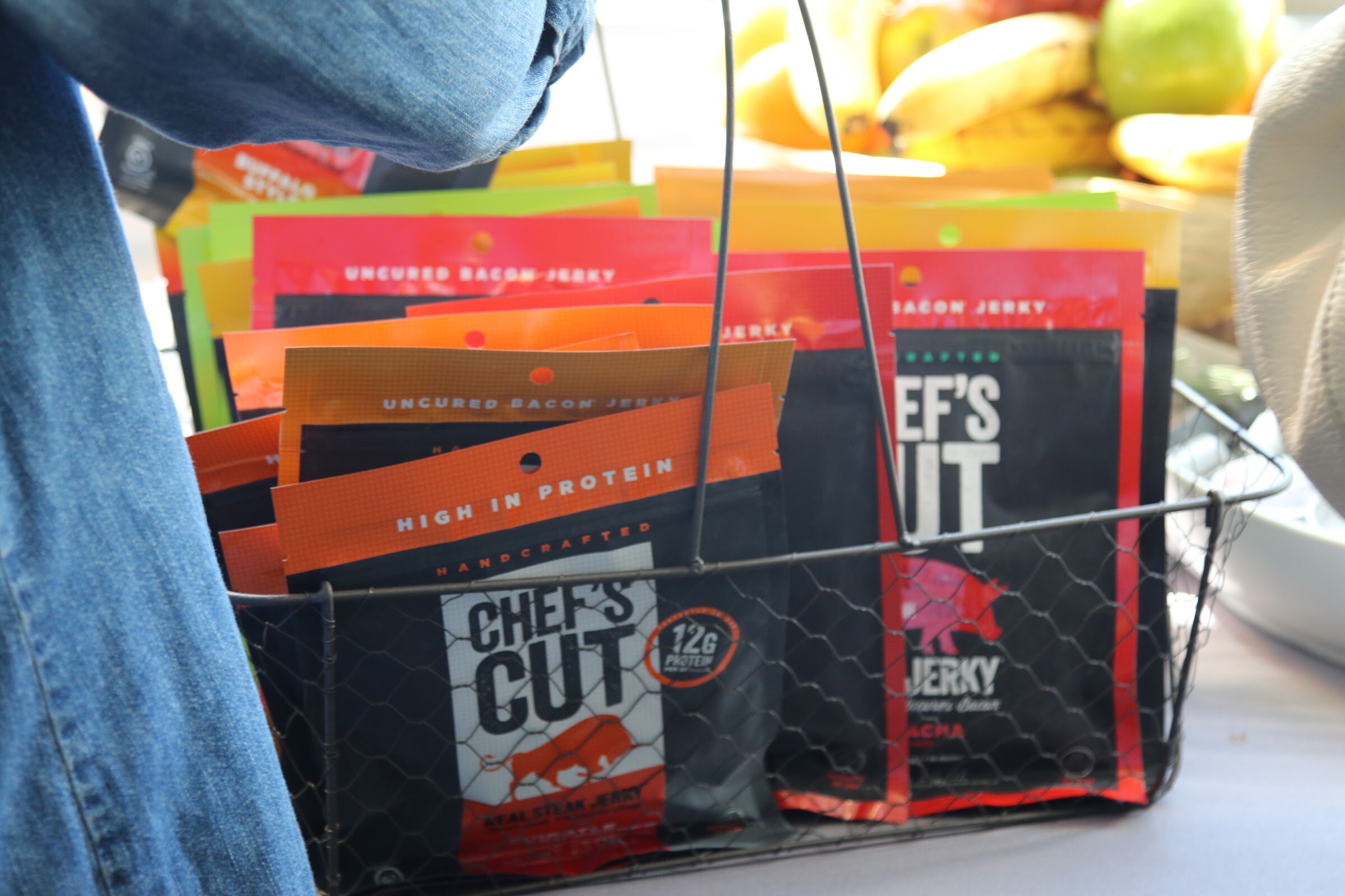 Wire basket of Chef's Cut bacon jerky bags in front of bananas and apples.