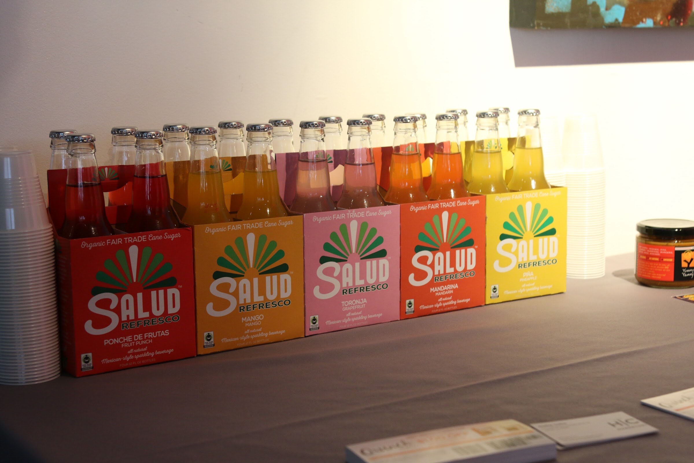 Salud Refresco Mexican-style sparkling beverage four-packs in five flavors with glass bottles on table.