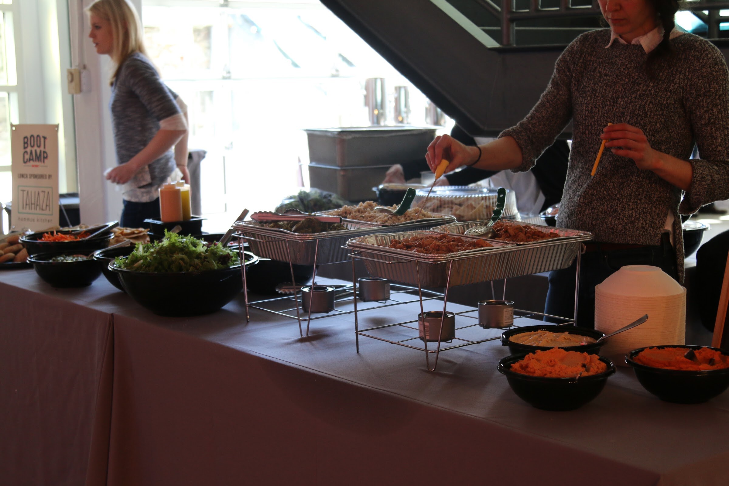 Lunch buffet table with chafing dishes, salad bowls, hummus, Boot Camp sign, servers.